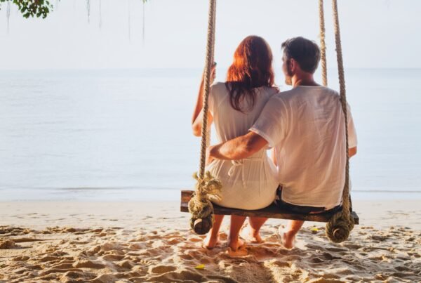 A couple sitting on a swing on a sandy beach, facing the calm sea at sunset.