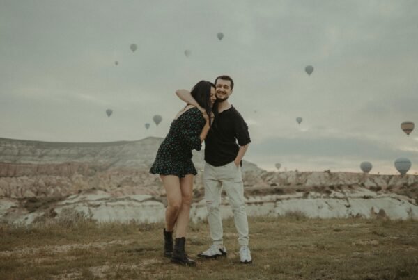 A couple kissing on a grassy area with hot air balloons floating in the sky.