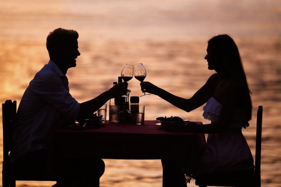 A couple toasting with wine glasses at a table by the sea during sunset.