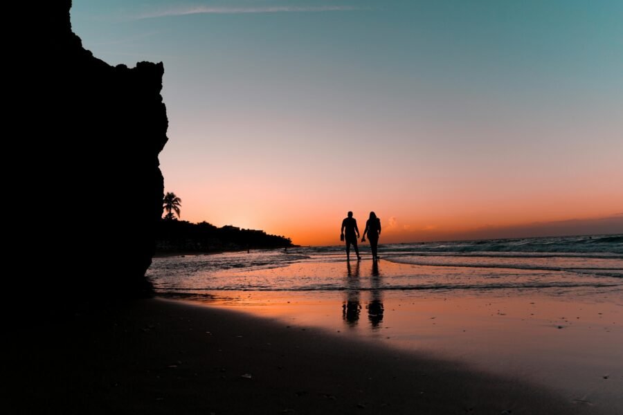 A couple walking along a wet beach at sunset, reflected in the shallow water.